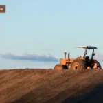 спецтехніка в оренду Трактор працює на широкому полі на світанку або заході сонця. Трактор знаходиться на вершині пагорба, підсвічений теплим світлом сонця, з небом і хмарами на задньому плані. Зображення підкреслює величину і простір сільськогосподарських робіт.