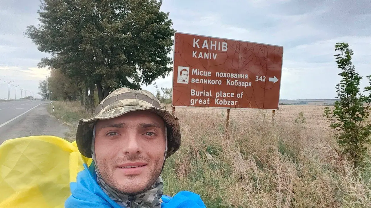 На зображенні показаний чоловік у військовій формі та кепці, який зробив селфі на тлі дорожнього знака, що вказує на напрямок до Канева — місця поховання великого Кобзаря, Тараса Шевченка. Напис на знаку дублюється українською та англійською мовами: "Місце поховання великого Кобзаря" та "Burial place of great Kobzar". Чоловік, імовірно, здійснює піший похід і на своєму маршруті проходить через важливі для української історії місця.