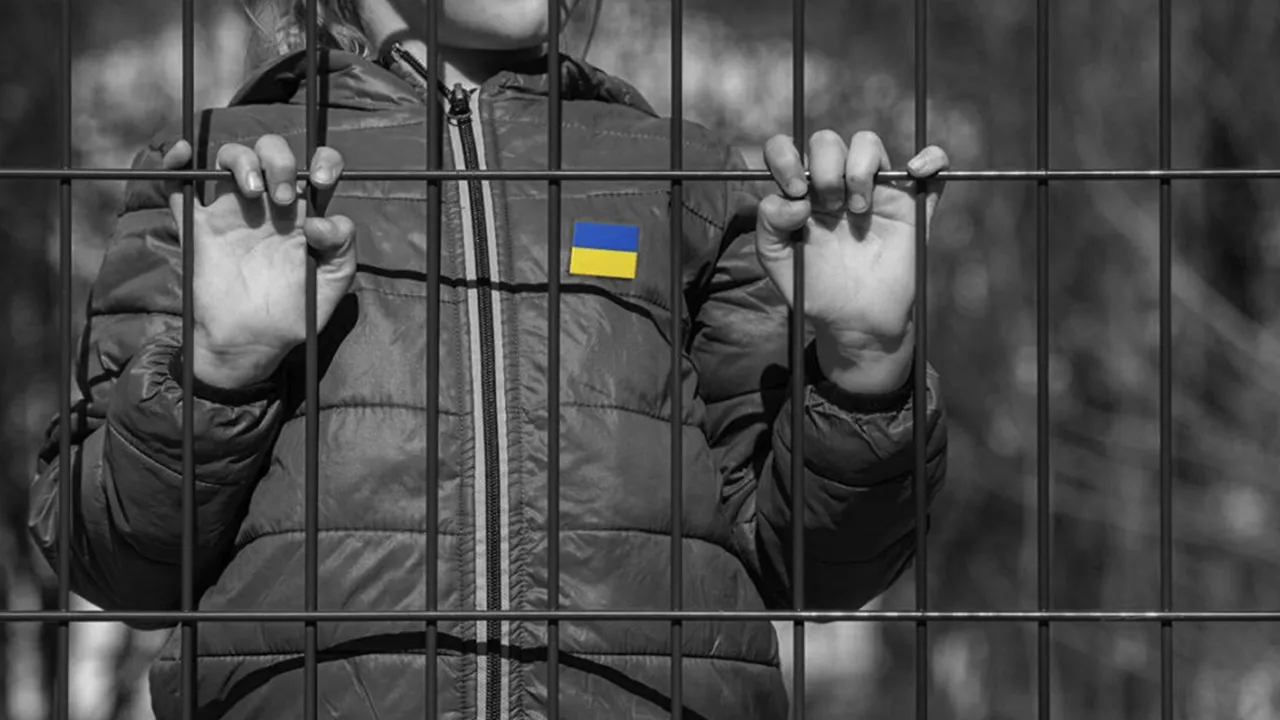На зображенні видно дитину в куртці, яка тримається за металеві ґрати обома руками. На куртці закріплено маленький прапор України. Фото створює враження замкнутості або обмеження свободи дитини, акцентуючи увагу на ситуації з дітьми в умовах конфлікту або окупації. Тло та більшість елементів зображені в чорно-білих тонах, що підсилює емоційний ефект, тоді як прапор має яскраві кольори, символізуючи надію або приналежність до України.