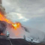 горить цистерна На фото зображені пожежники в спеціальних захисних костюмах, які гасять палаючу залізничну цистерну. Полум'я виривається з верхньої частини цистерни, піднімаючись високо в небо. Видно густий чорний дим, що піднімається у небо, та піну, яку пожежники використовують для гасіння вогню.