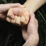 hands На зображенні видно дві пари рук. Одна рука міцно тримає зап'ястя іншої, обидві руки виглядають напруженими. Довкола рук видно зелень, що свідчить про те, що дія відбувається на відкритому повітрі, можливо, серед трави.