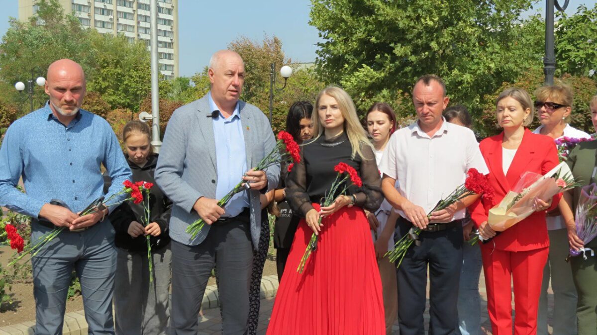 Група дорослих стоїть з квітами на площі під час акції. У центрі стоїть чоловік у світло-сірому піджаку, поряд із ним – жінка в чорному топі та червоній спідниці. Всі учасники мають серйозний вираз обличчя, багато хто тримає червоні гвоздики, які, ймовірно, будуть покладені до меморіалу.