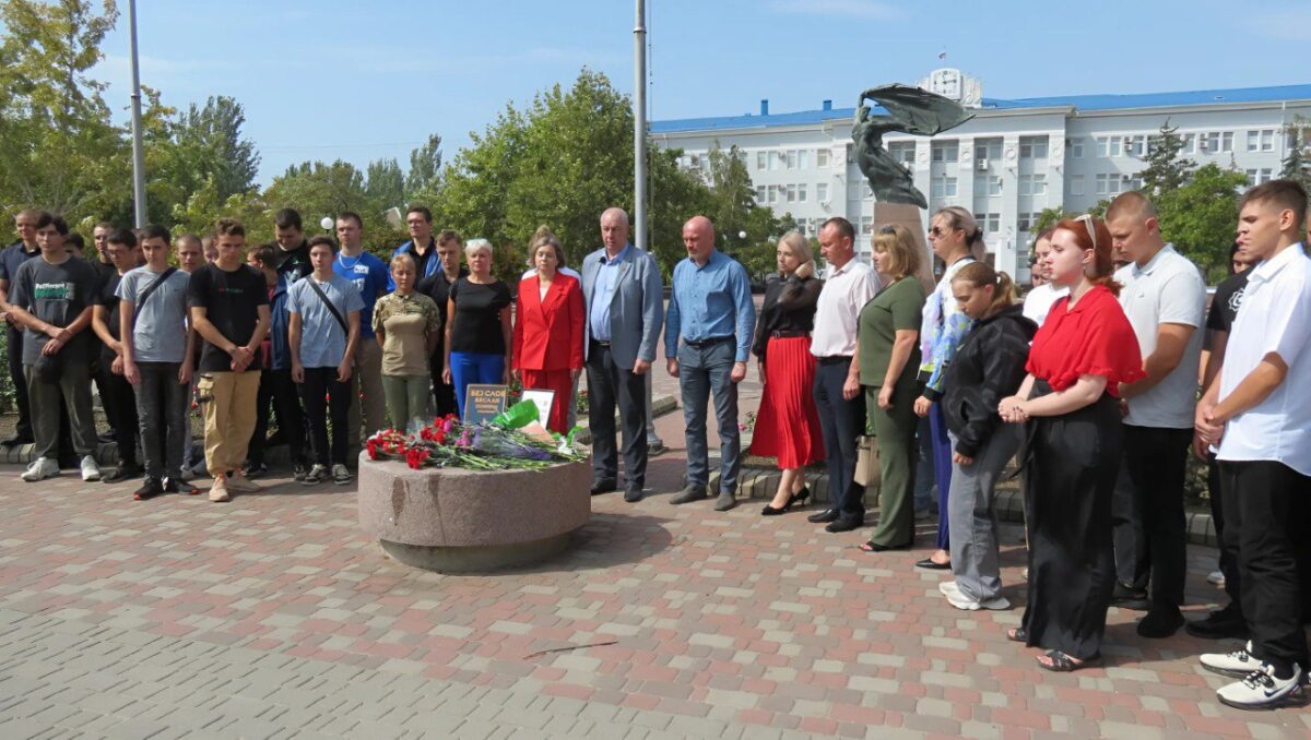 Група людей зібралася на площі біля меморіалу для проведення пам'ятної акції. На передньому плані видно молодь, ймовірно, учасників з місцевих молодіжних організацій, та кількох дорослих, які стоять поруч. Усі вони мають серйозний вигляд і тримають руки складеними перед собою або за спиною. Попереду меморіалу лежать квіти, які, ймовірно, були покладені учасниками акції.